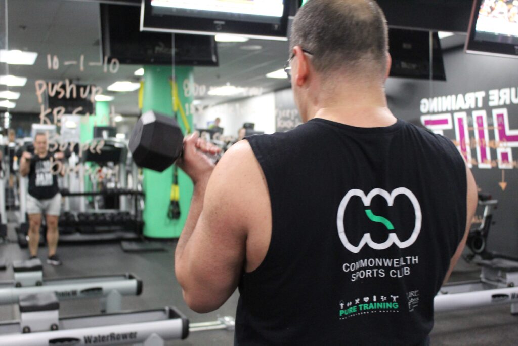 CSC member lifting dumbbells in front of a mirror; you see his back and the member is wearing a CSC branded tank top with the CSC logo.